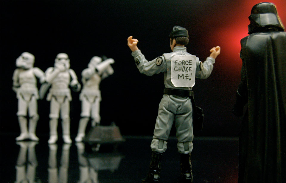 A photo of Star Wars toys showing Stormtroopers laughing at a Death Star worker who has a sign on his back saying "Force Choke Me" with Darth Vader looking on.