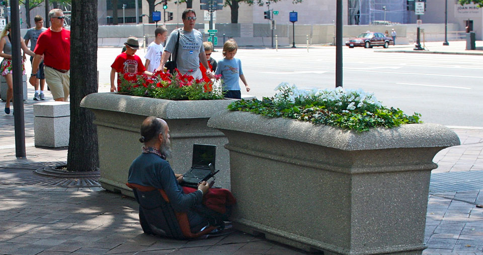 A photo of an old man with a pony tail sitting in the street with a laptop open.