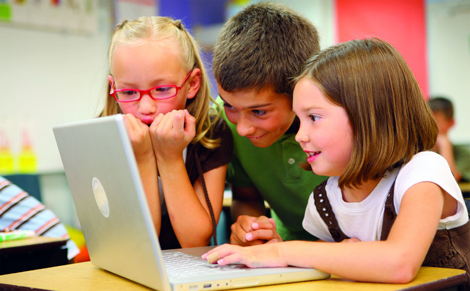 A photo of three children smiling and grouped around a laptop.