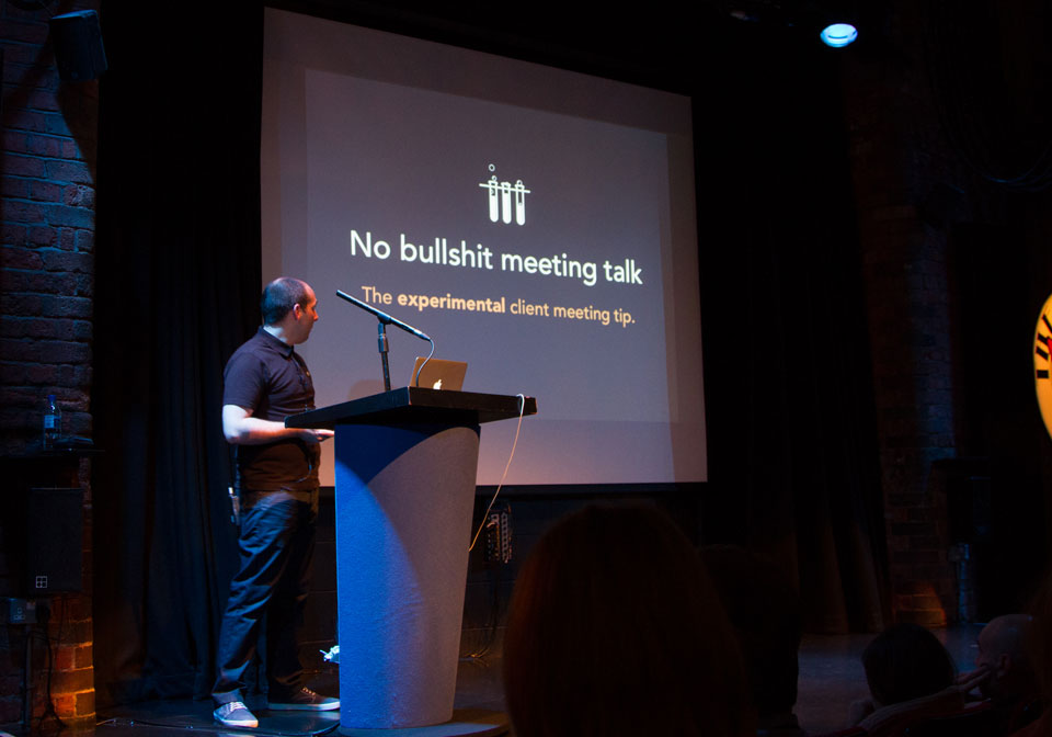 A photo of Sam on stage behind a lectern in front of one of his slides that says No bullshit meeting talk.