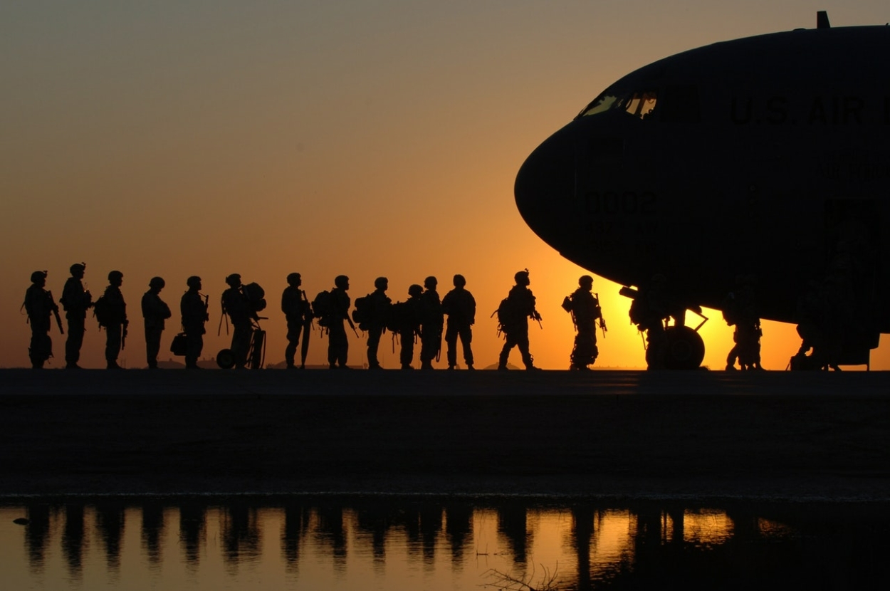A photo taken during an orange sunset, showing the silhouettes of soldiers, lining up to board a plane, which is seen on the right-hand side of the picture.