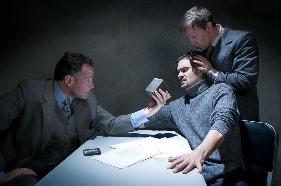 A photo of a staged interrogation scene with one man holding a box to the face of a man while another holds the head of the man who looks distressed