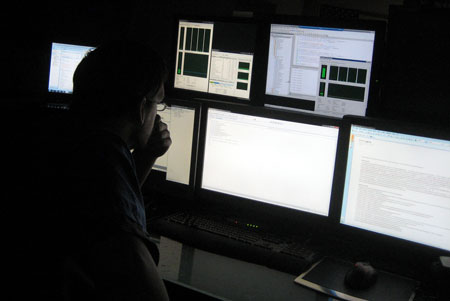 A man sitting in the dark in front of six computer monitors