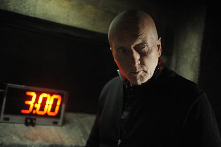 A movie screenshot of Jigsaw with a menacing stare in front of a countdown clock